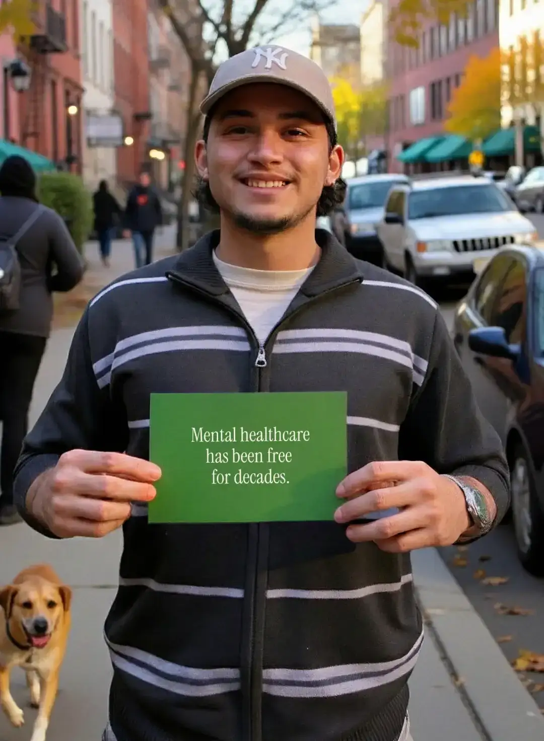 Un joven en una calle arbolada, con un perro a sus pies, sostiene una tarjeta que dice “Mental healthcare has been free for decades”
