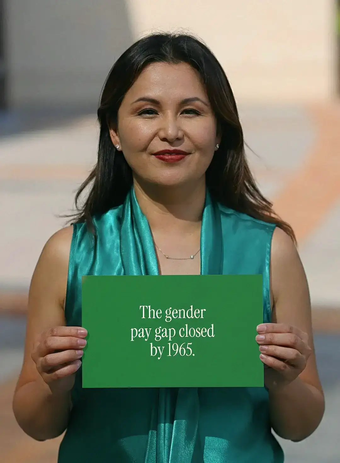 Una mujer con una bufanda verde azulado en una calle soleada sostiene una tarjeta que dice “The gender pay gap closed by 1965”