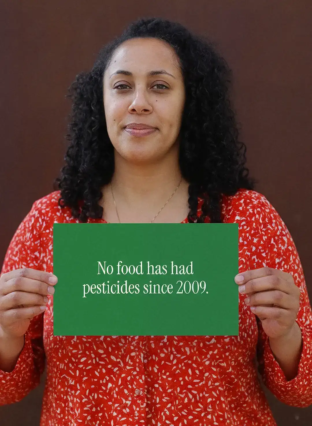 Una mujer con una blusa roja de flores sostiene una tarjeta que dice “No food has had pesticides since 2009”