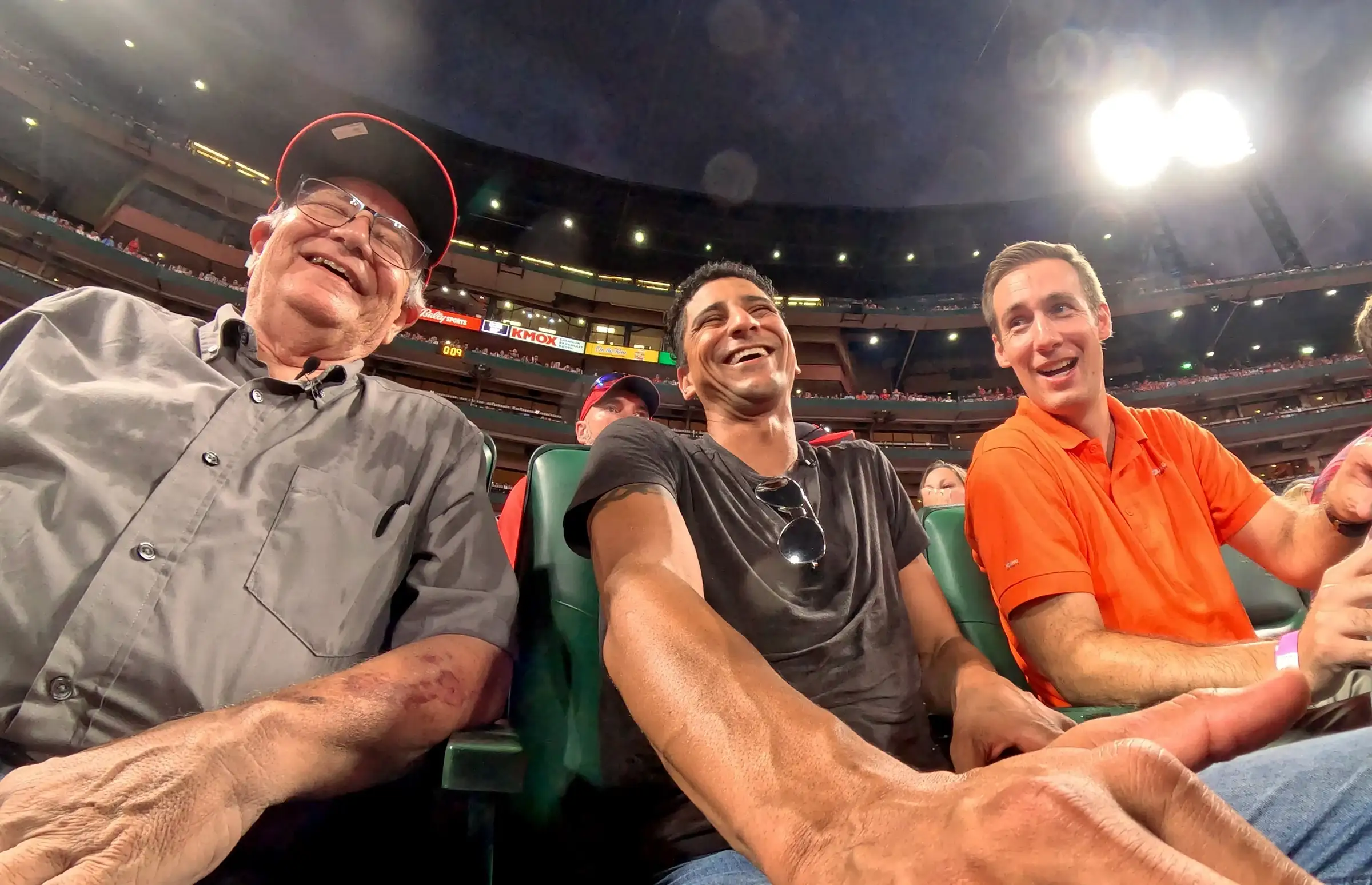Jared Young, con una camiseta polo naranja, sentado junto a John y Julian en las gradas del Busch Stadium mientras los tres se ríen
