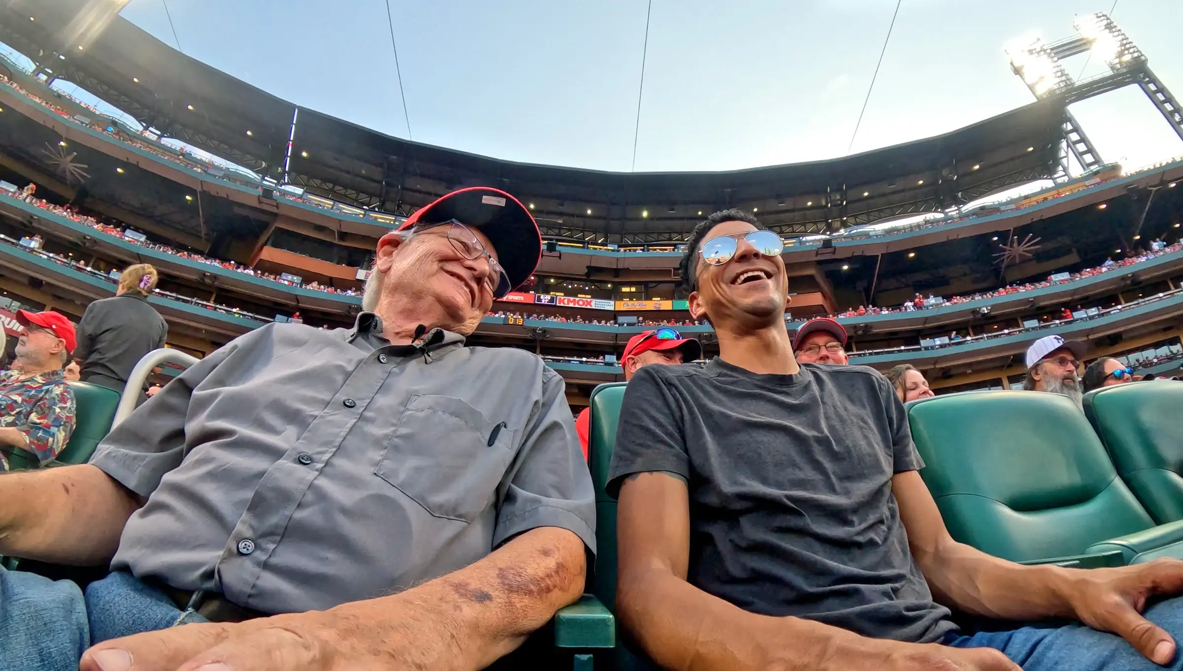 Un hombre mayor con gorra de los Cardinals y un hombre más joven con gafas de sol se ríen juntos en asientos verdes detrás del home en el Busch Stadium