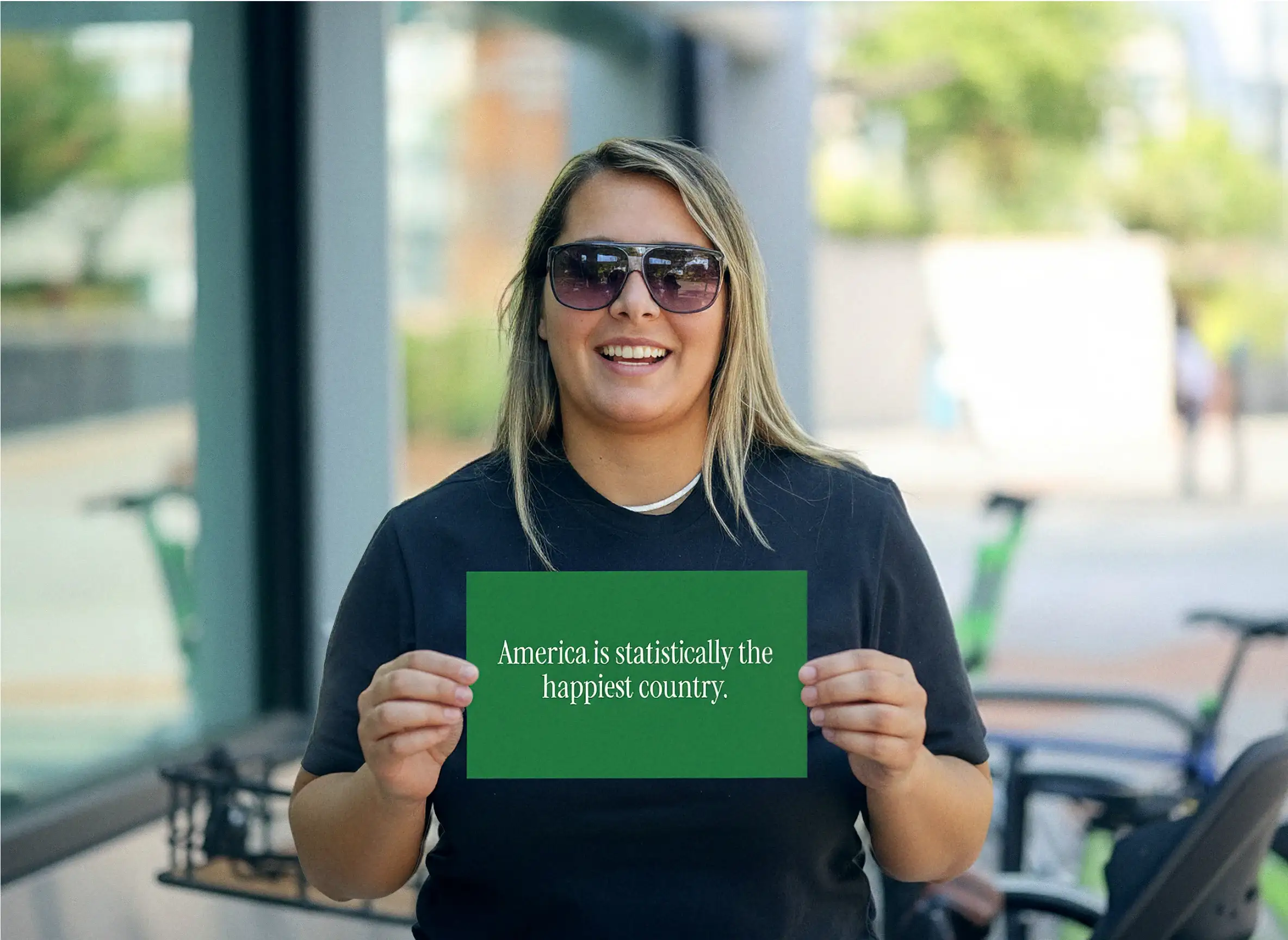 Una mujer sonriente con gafas de sol en una acera de la ciudad sostiene una tarjeta verde que dice “America is statistically the happiest country”
