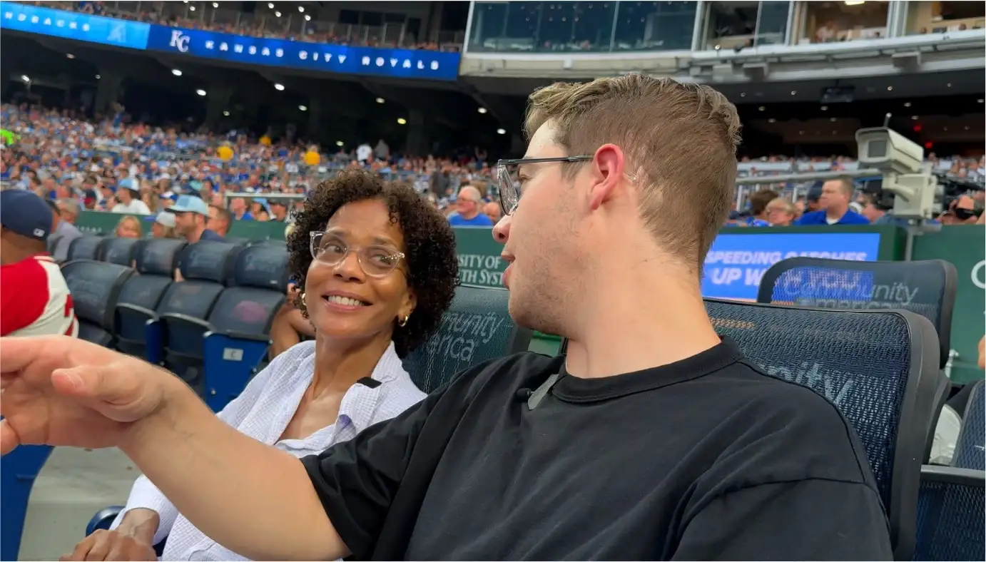 Kirby, con una blusa blanca, y Luke, con una camiseta negra, conversan en sus asientos del Kauffman Stadium