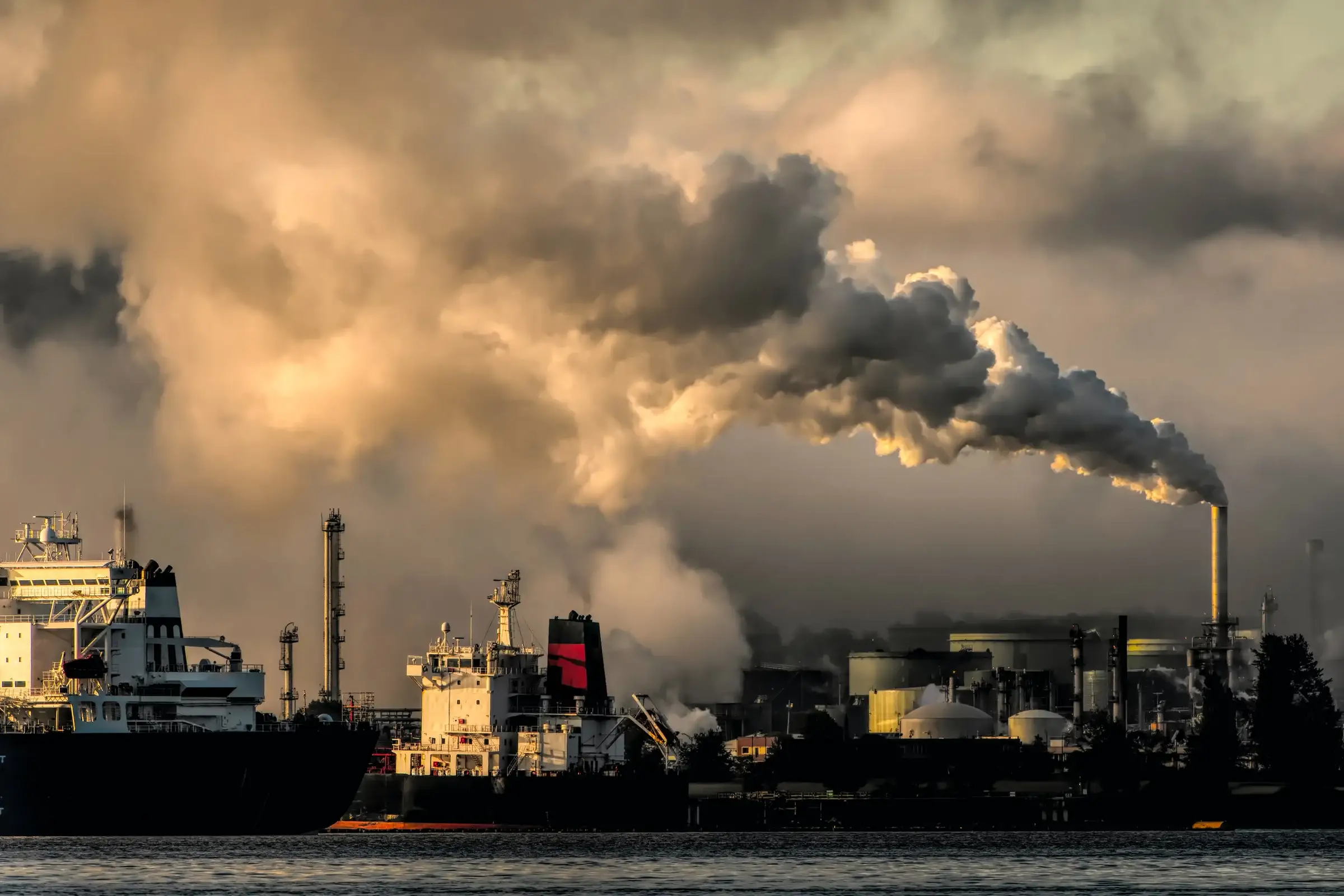 Smokestacks pour grey smoke into a cloudy sky over a harbor, with ships in the foreground