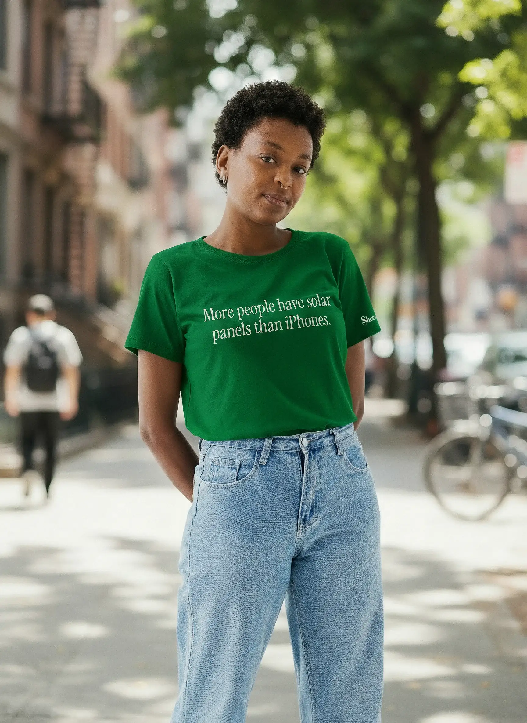 Una mujer en una acera de la ciudad lleva una camiseta verde que dice “More people have solar panels than iPhones”
